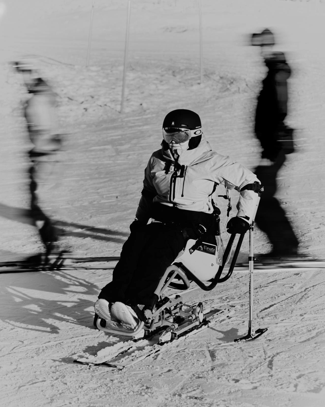 Black and white photo of a lady in a sit ski with blurred skiers in the background